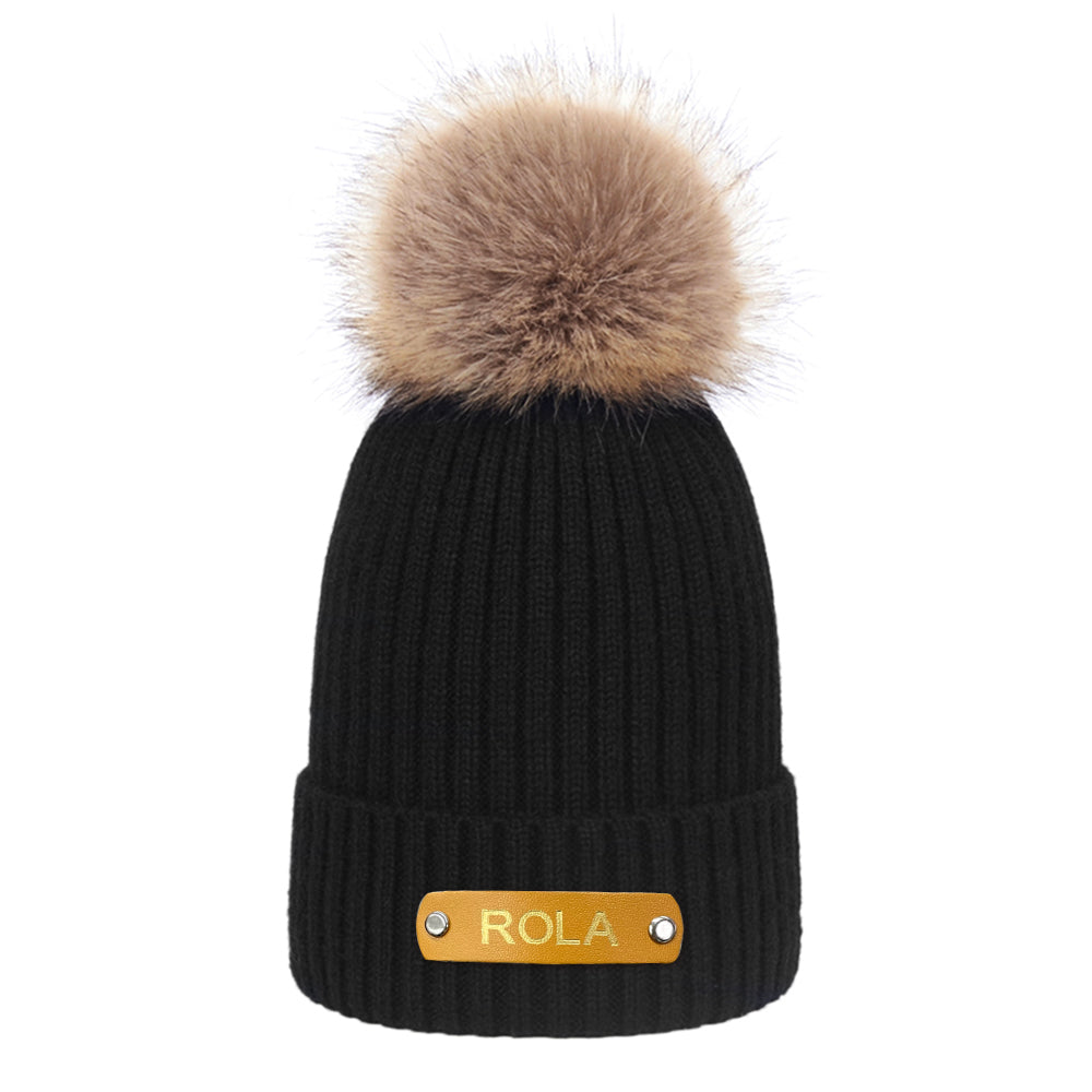 Black knit beanie with a fur pom-pom and gold 'ROLA' label on a white background