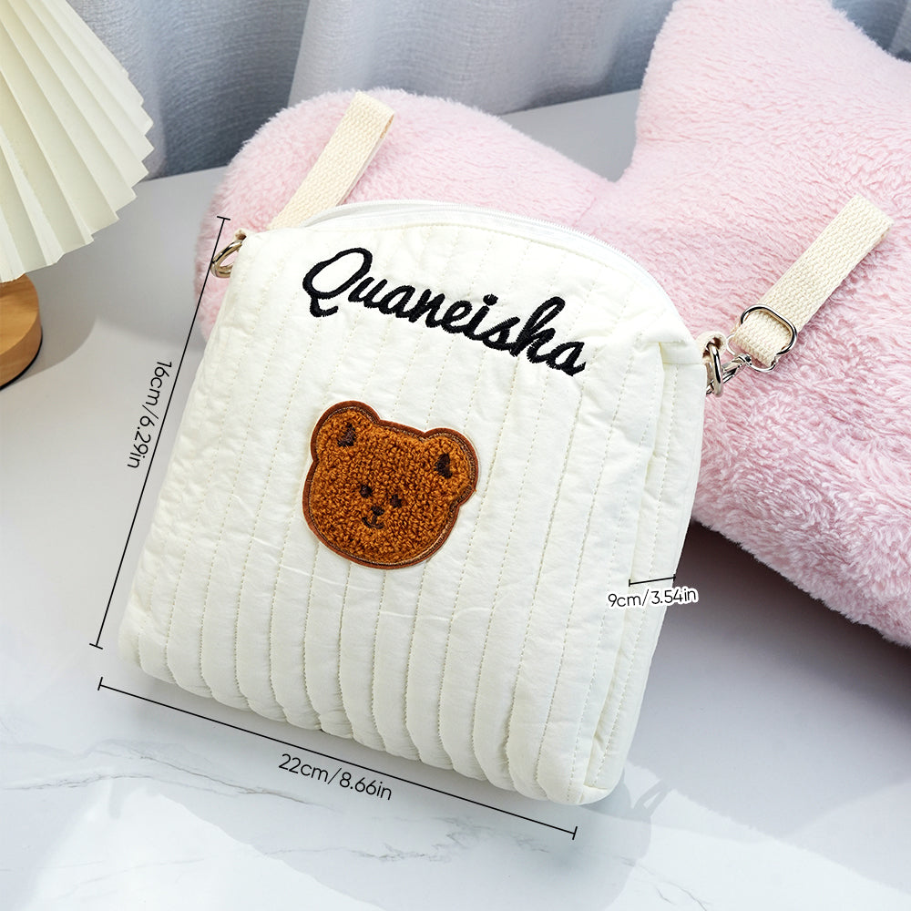 Small white backpack with teddy bear emblem and 'Quaneisha' text, placed on a light surface with pink plush toy in the background.