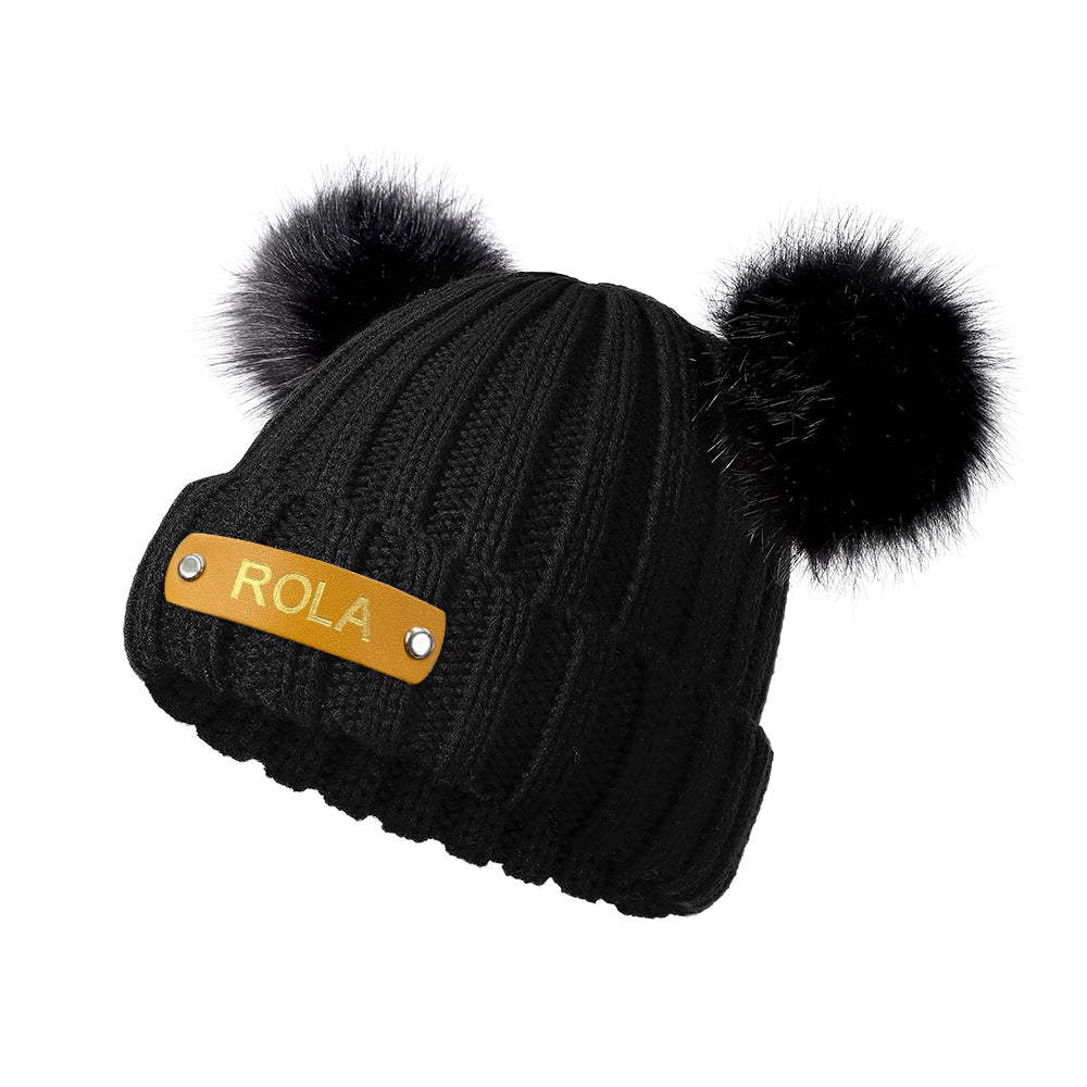 Black beanie with two pom-poms and a gold label on a white background