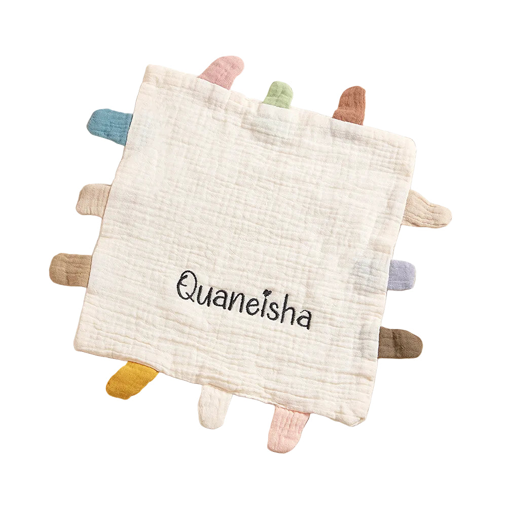 Baby comfort blanket with colorful tassels and 'Quaneisha' branding on a soft beige background