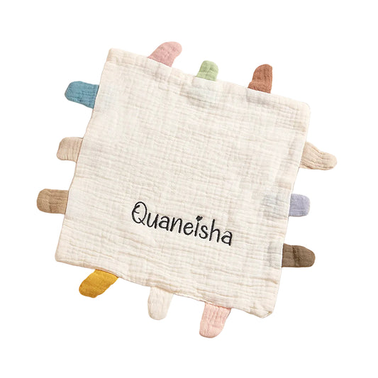 Baby comfort blanket with colorful tassels and 'Quaneisha' branding on a soft beige background