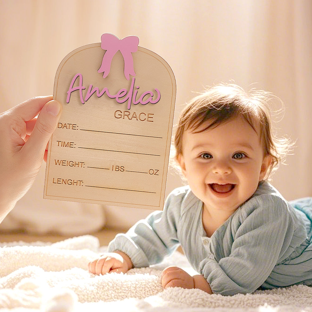 Baby lying on a blanket with a wooden sign featuring the name 'Amelia Grace' held by an adult.