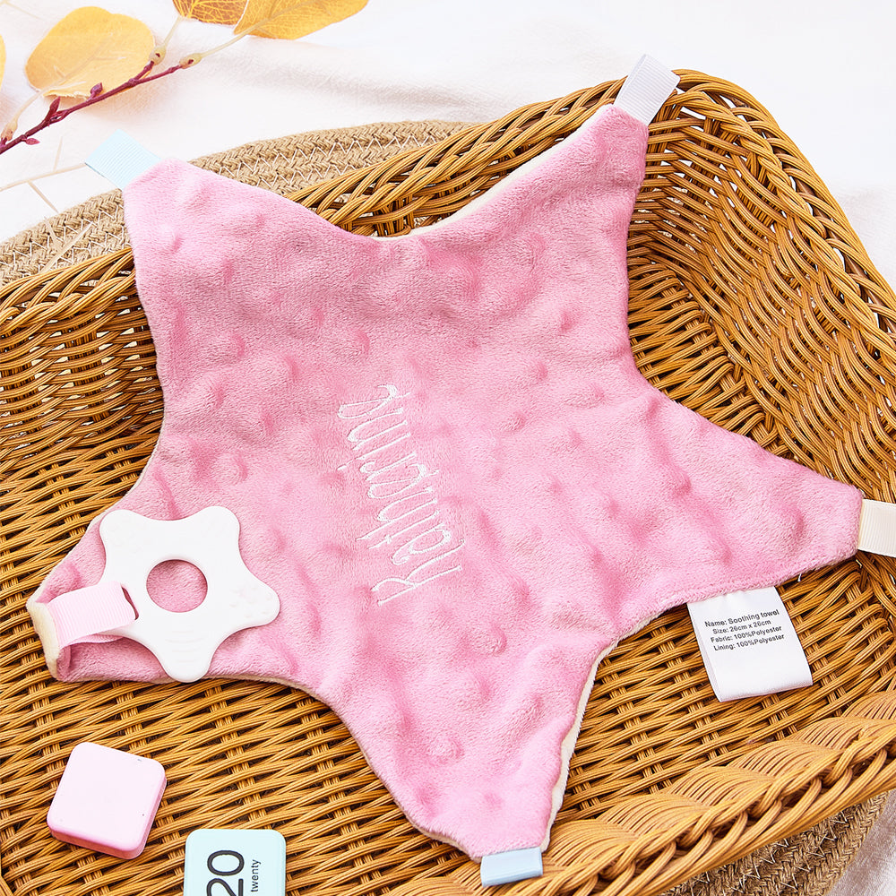 Pink star-shaped baby comfort blanket with a white teether on a wicker surface