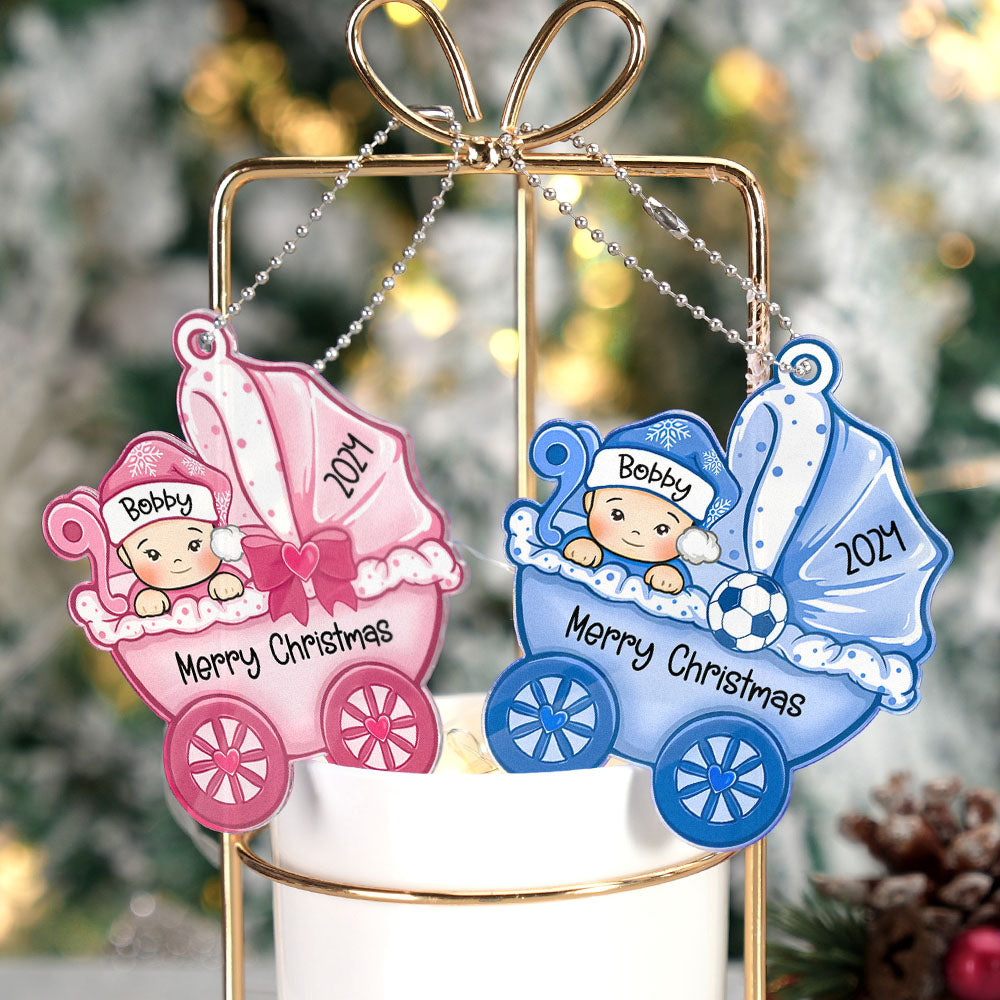 Two Christmas ornaments shaped like baby carriages, one pink and one blue, with personalized names and 'Merry Christmas' text, against a blurred Christmas tree background.