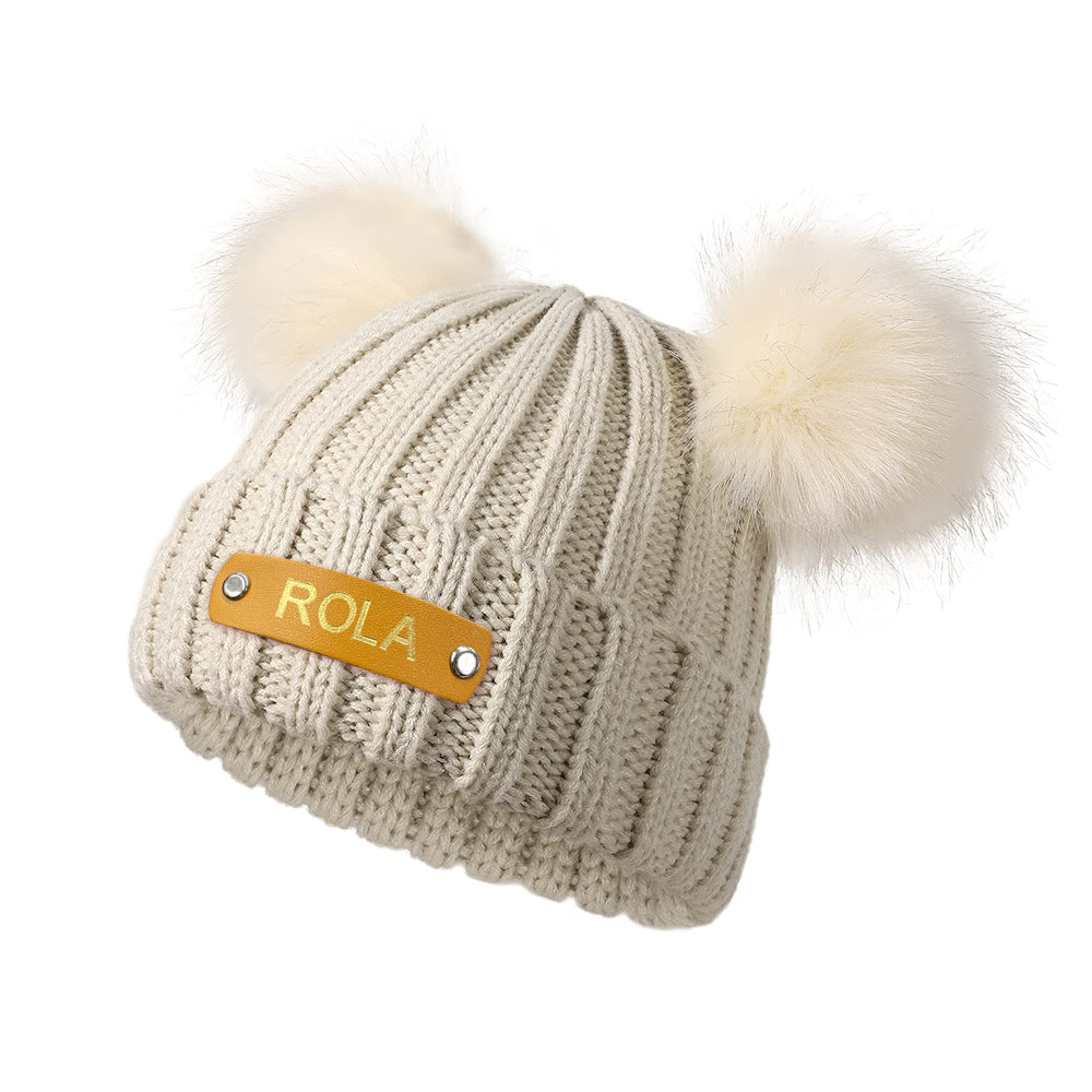 Beige knit beanie with white pom-poms and a gold patch labeled 'Rola' on a white background