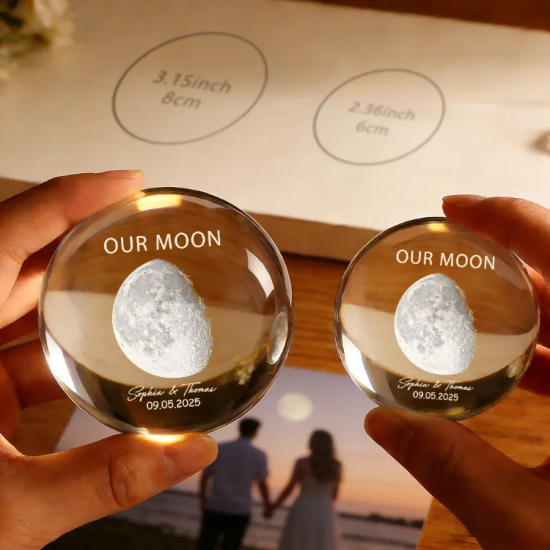 Two hands holding clear crystal balls with engraved 'Our Moon' and personalized names, set against a blurred background of a couple holding hands.