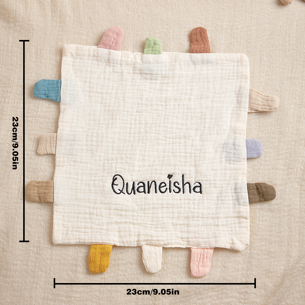 Baby comfort blanket with colorful tabs and 'Quaneisha' branding, displayed on a textured surface with measurements.
