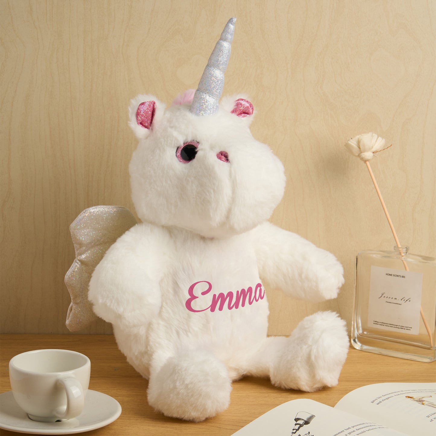 White teddy bear with unicorn horns and ears, sitting on a wooden surface with a cup and book in the background.