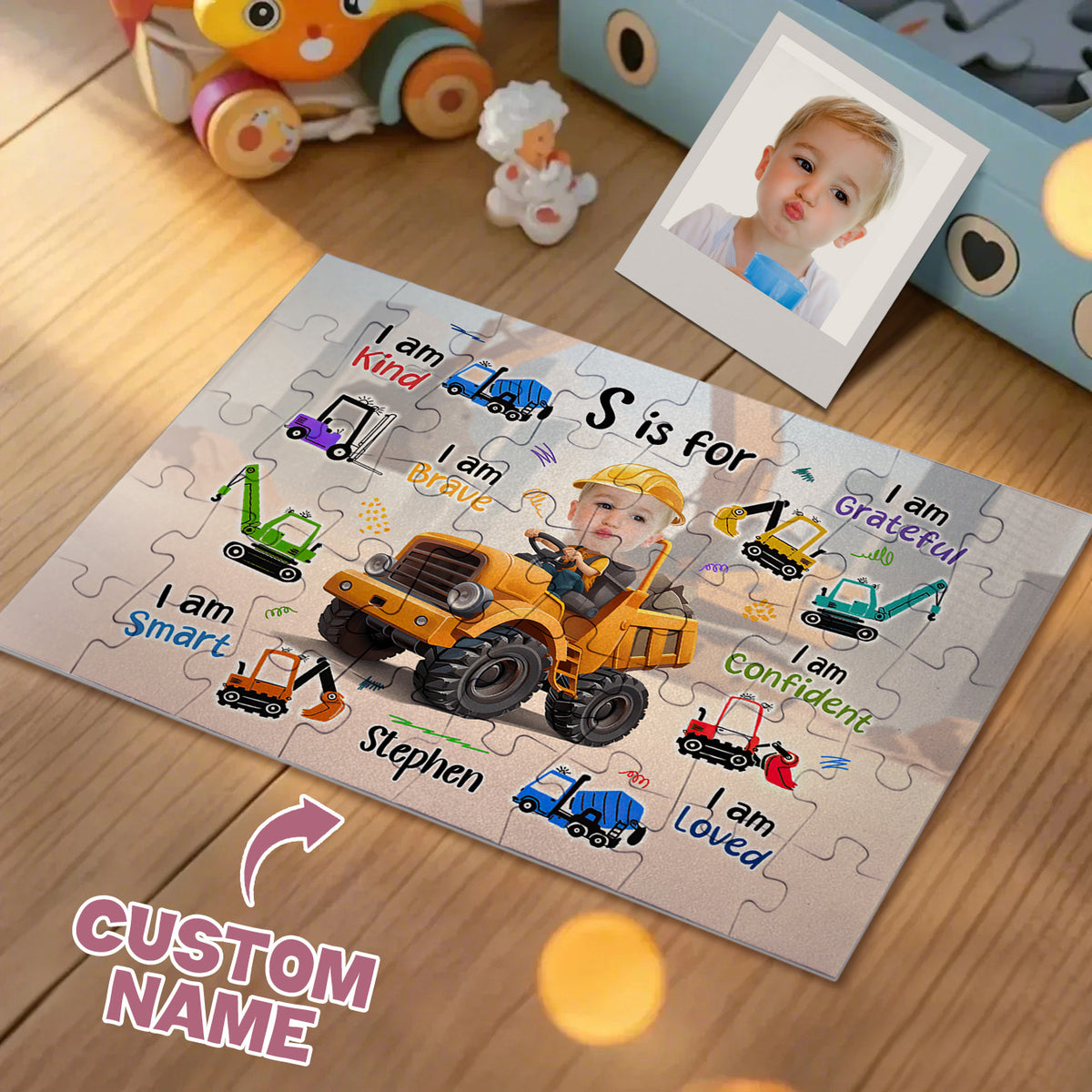 A custom children's jigsaw puzzle displayed on a wooden floor, featuring a photo of a baby in a yellow construction hat driving a yellow excavator, surrounded by customizable text like 'I am Kind', 'I am Brave', and 'I am confident'. A photo frame with a baby's portrait and toys are arranged nearby on the floor.