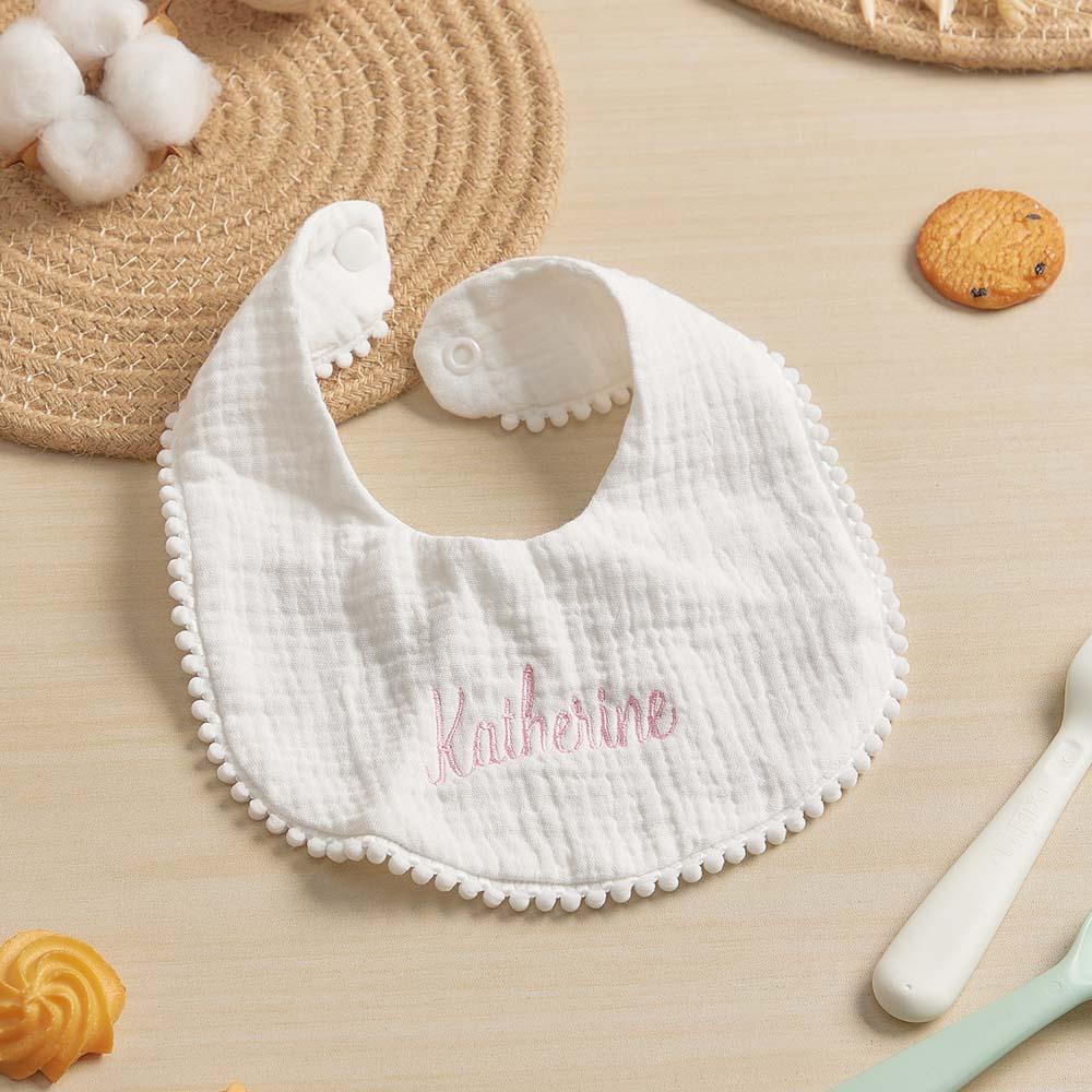 White baby bib with 'Katherine' embroidery on a beige surface with cookies and a straw hat.