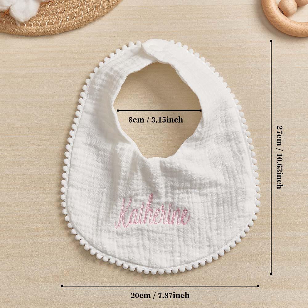 White baby bib with pom-pom trim and embroidered name, shown with measurements on a beige surface.