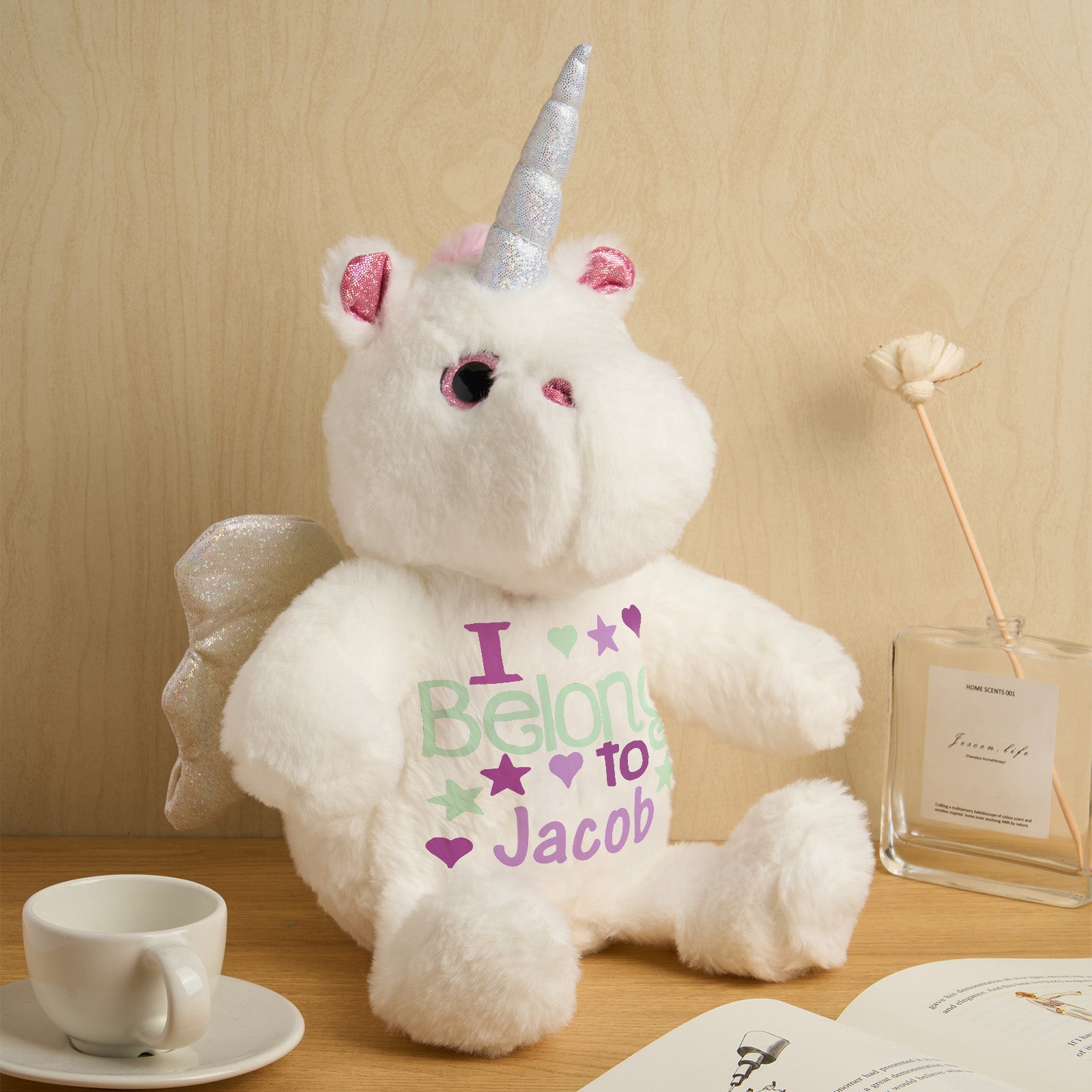 White teddy bear with unicorn horn and wings wearing a shirt that says 'I Belong to Jacob' on a wooden surface.