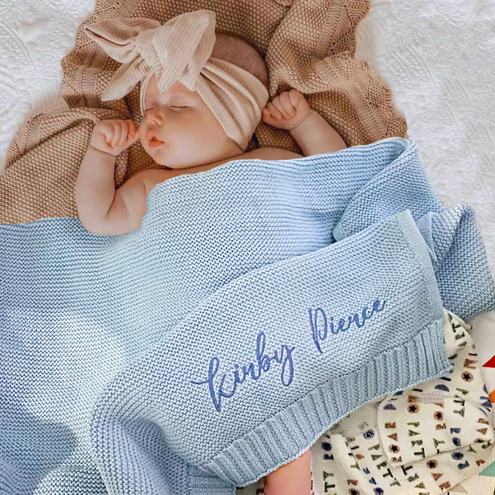 Newborn baby wrapped in a blue blanket with embroidered name, wearing a beige headband, on a soft surface.