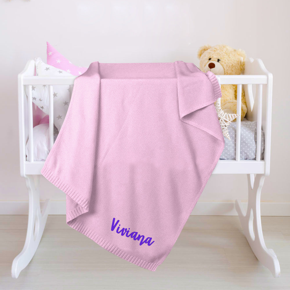 Pink baby blanket with 'Viviana' embroidery on a white crib with a teddy bear and baby shoes.