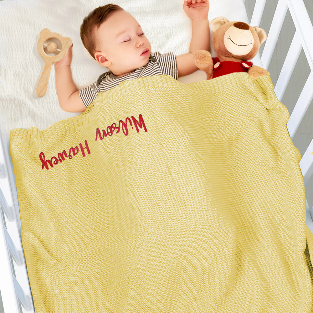 Baby lying in a crib with a yellow blanket and toy, wearing a striped shirt.