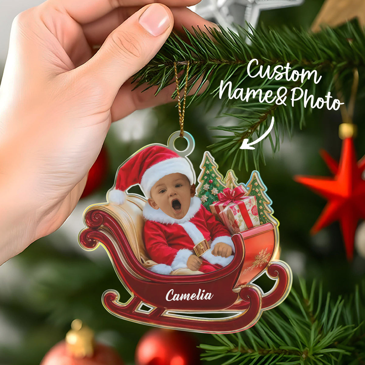 Christmas ornament with a baby in a sleigh, held by a hand on a decorated tree.