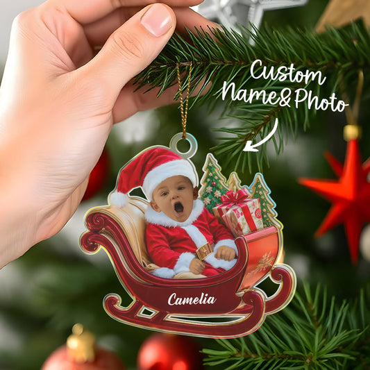 Christmas ornament with a baby in a sleigh, held by a hand on a decorated tree.