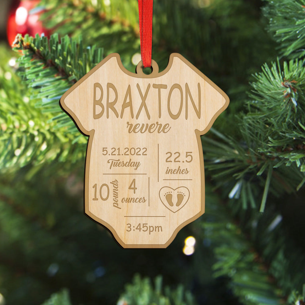 A personalized baby shower baby clothes ornament shaped like a onesie, hanging on a Christmas tree. The natural wood ornament is engraved with the name 'BRAXTON', the word 'revere', and baby birth details, and hangs from a red ribbon against a green pine tree background.