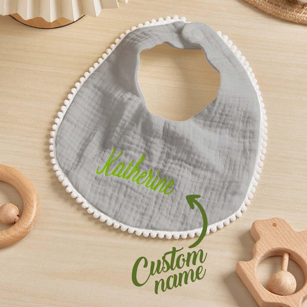 Gray baby bib with white trim and embroidered name on a wooden surface with wooden toys.
