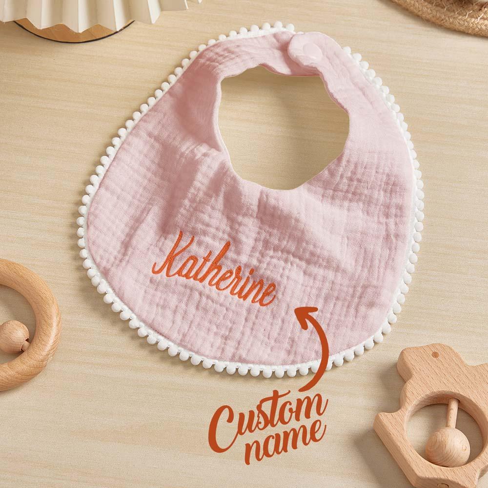 Pink baby bib with embroidered name on a wooden surface with toys around