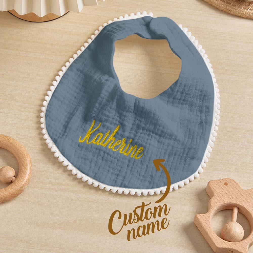Blue baby bib with embroidered name on a wooden surface with toys.