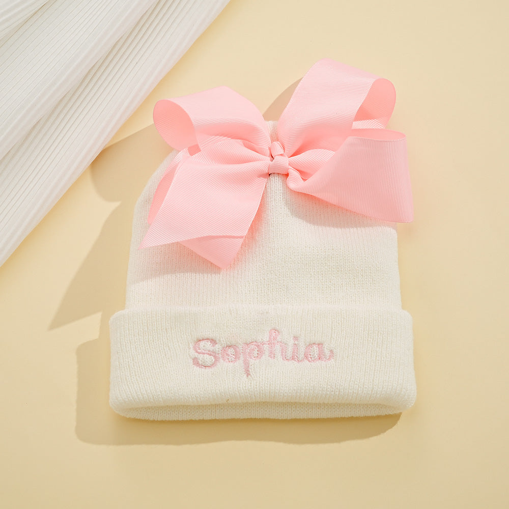White baby hat with pink bow and embroidered name 'Sophia' on a beige background
