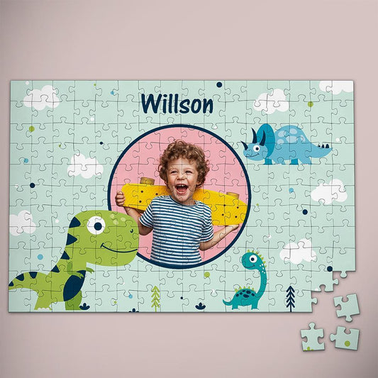 A completed personalized children's jigsaw puzzle on a light pink background. The puzzle features cartoon dinosaurs, white clouds, and the name 'Willson' at the top, with a circular cutout containing a child's photo.