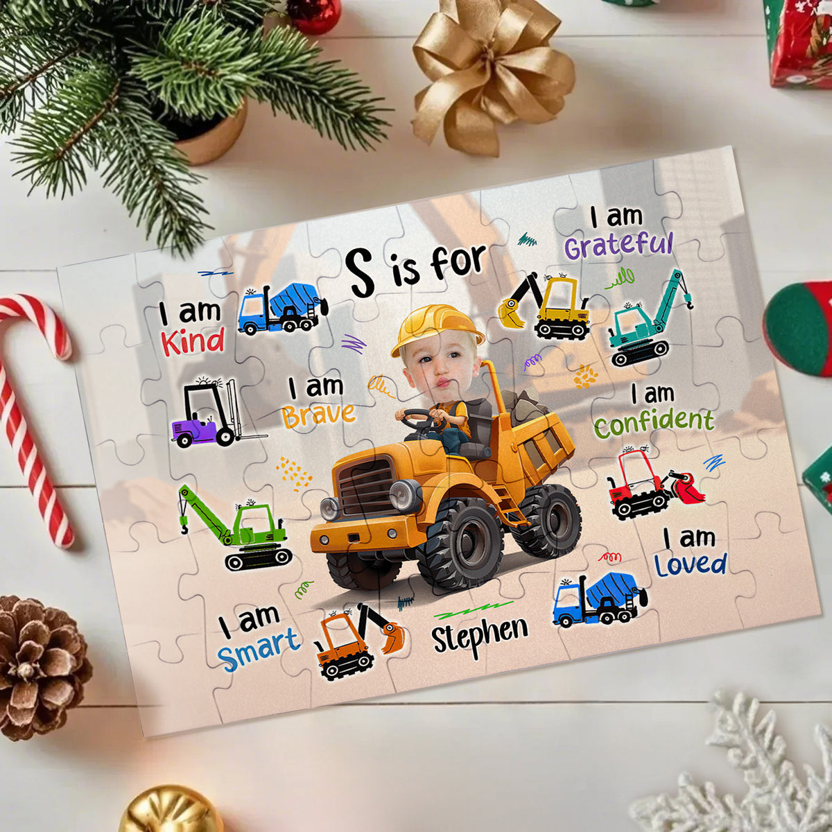 A personalized jigsaw puzzle featuring a baby construction worker driving a yellow dump truck, surrounded by construction vehicles and positive phrases like 'I am Kind', 'I am Brave', and 'I am Confident'. The puzzle is styled on a white surface with Christmas decorations including a candy cane, pinecone, greenery, and a gold bow.