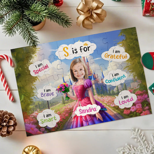 A personalized jigsaw puzzle featuring a princess girl in a pink dress standing in a colorful garden with the text 'S is for' and positive affirmations like 'I am Grateful' and 'I am Smart.' The puzzle is styled in a holiday flat-lay setting with a gold bow, pine branches, candy canes, and a small ornament on a white wooden surface.