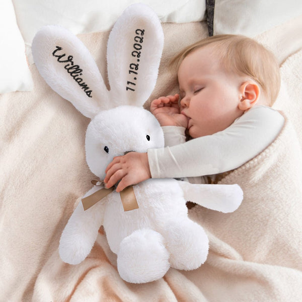 Baby holding a white plush bunny with personalized text, lying on a soft surface.