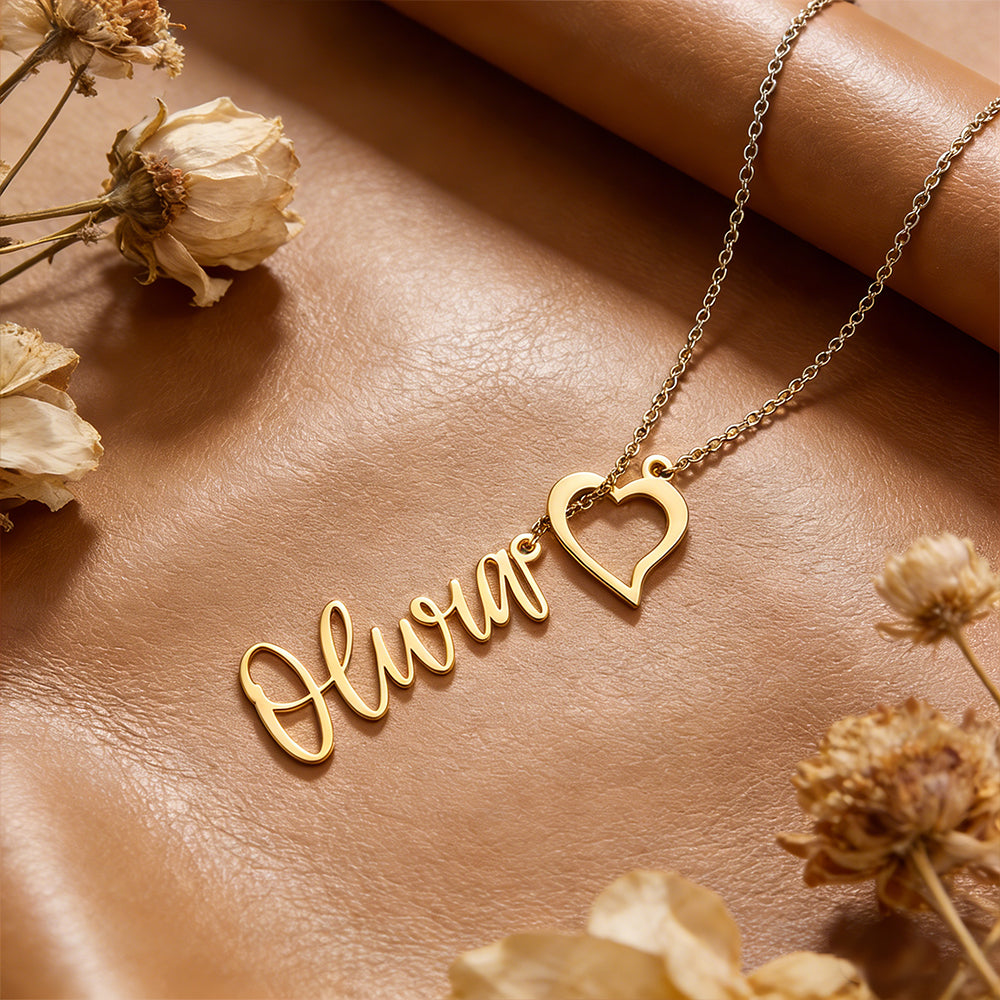 Gold necklace with 'Oliver' and heart design on a brown surface with dried flowers.
