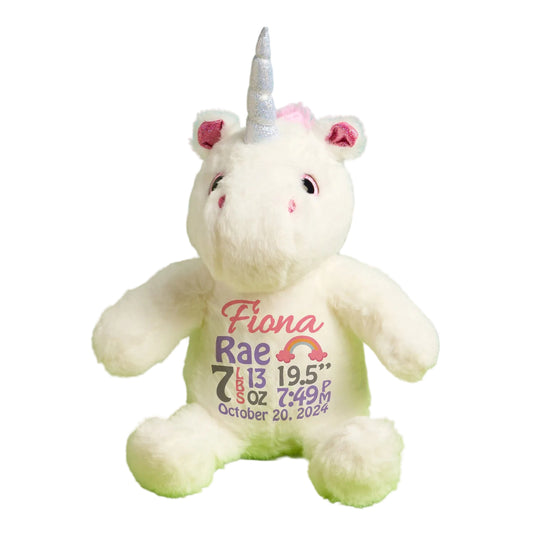 White unicorn plush toy with personalized text on a green surface, flowers, and plant in the background.