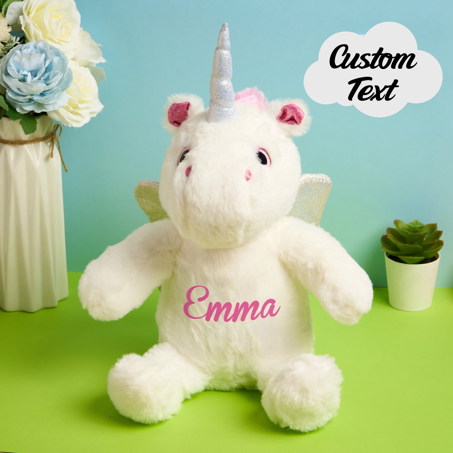 White unicorn plush toy with pink name on a green surface, flowers, and plant in the background