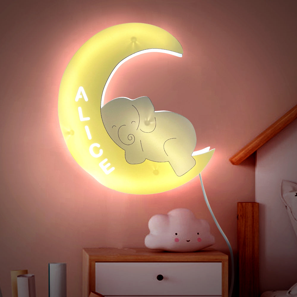 Moon-shaped night light with an elephant design and 'ALICE' text, placed on a shelf against a pink wall.