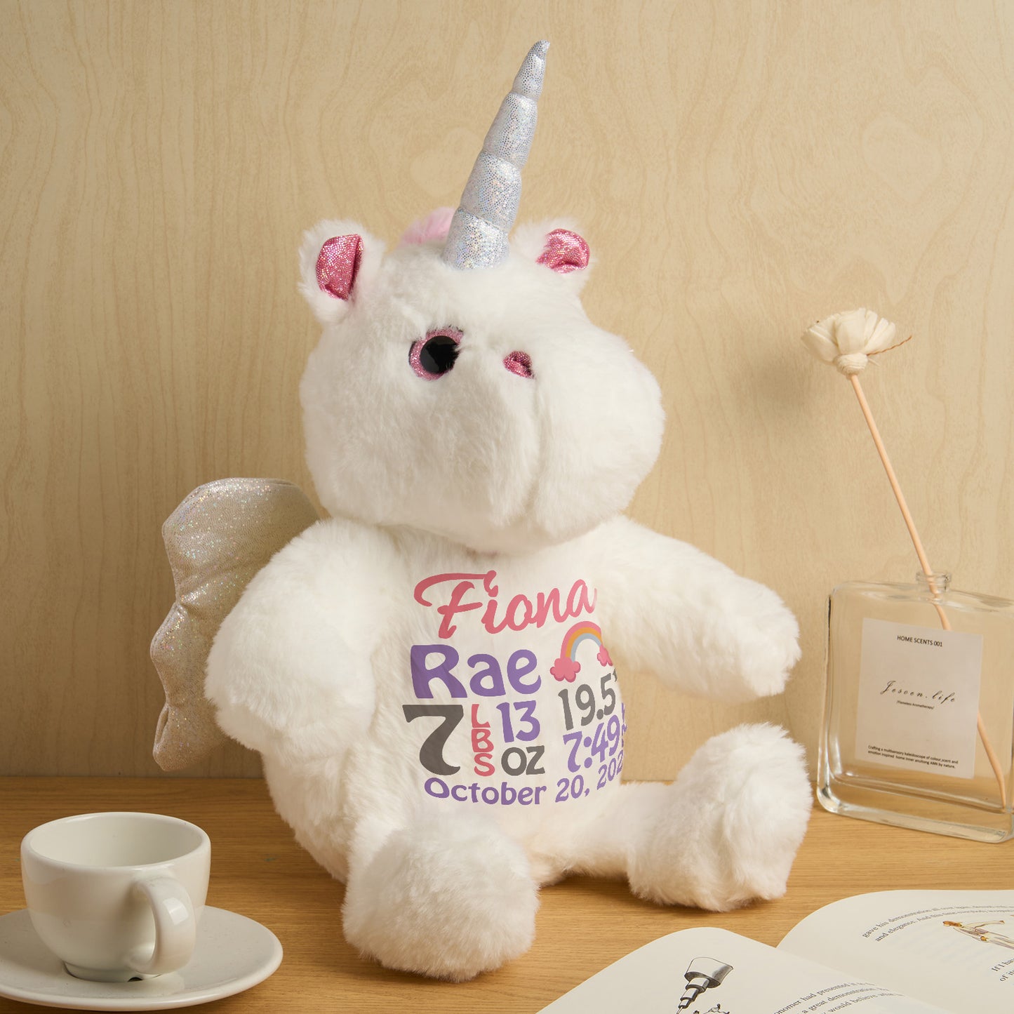 White teddy bear with unicorn horn and ears wearing a shirt with text, sitting on a wooden surface with a cup and plant in the background.