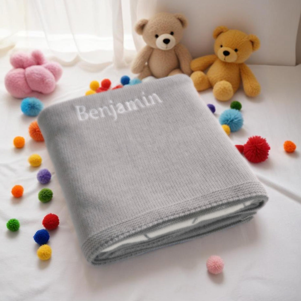 Gray blanket with 'Benjamin' embroidered, surrounded by colorful pom-poms and two teddy bears on a white surface.