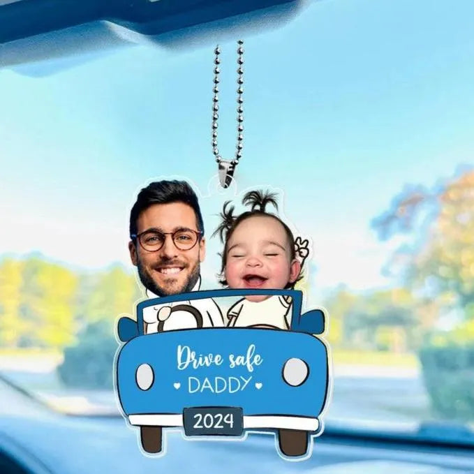 Car rearview mirror hanging with a cartoon design of a man and child in a car, text 'Drive safe DADDY 2024', and blurred outdoor scenery.