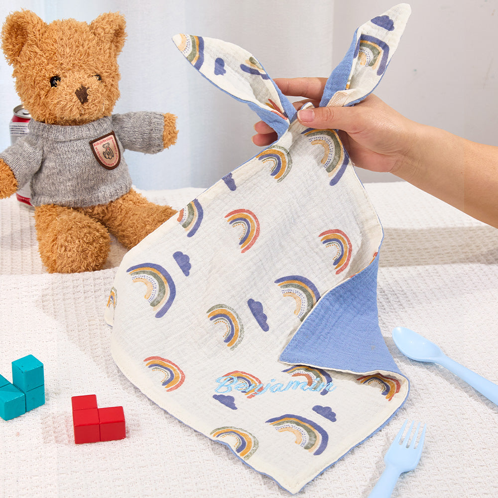 Baby blanket with rainbow pattern held by a hand, next to a teddy bear and toys on a white surface.