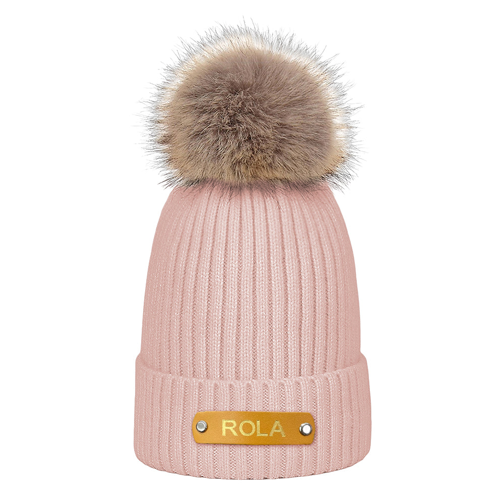 Pink knit beanie with fur pom-pom and 'Rola' label on a white background