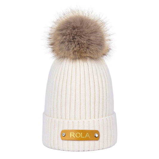 White knit beanie with brown fur pom-pom and gold 'Rola' tag on a white background