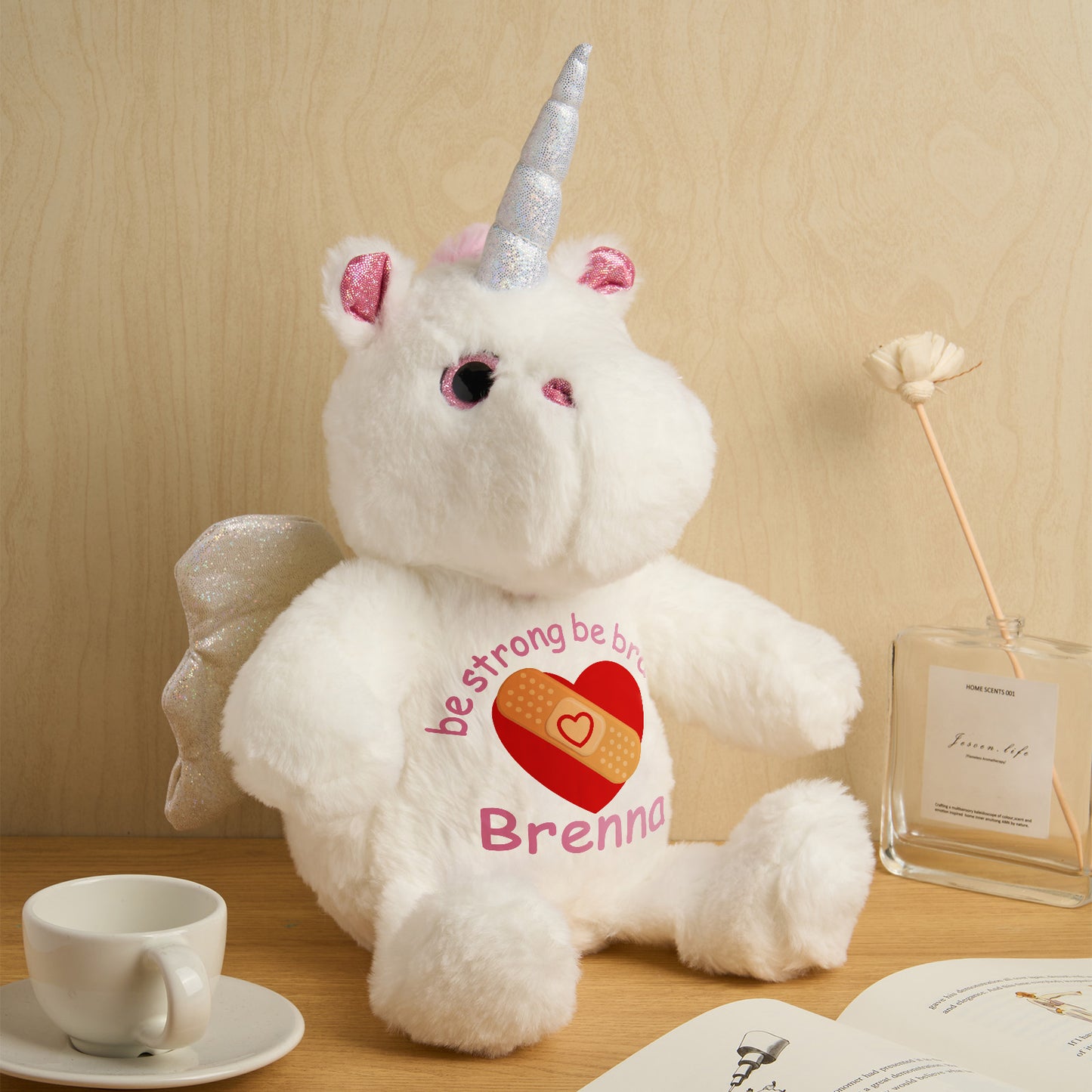 White teddy bear with unicorn horn and wings wearing a shirt with a heart and text, sitting on a wooden surface.