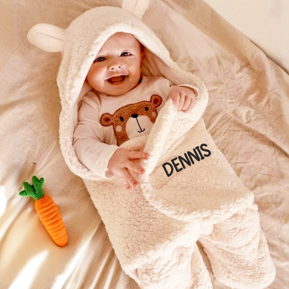 Baby wrapped in a beige hooded blanket with 'DENNIS' on it, smiling and holding a carrot toy.