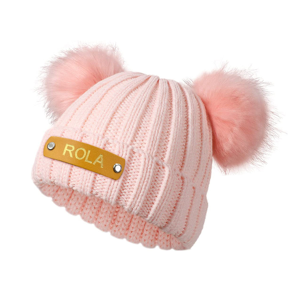 Pink knit beanie with two pom-poms and a gold tag labeled 'ROLA' on a white background